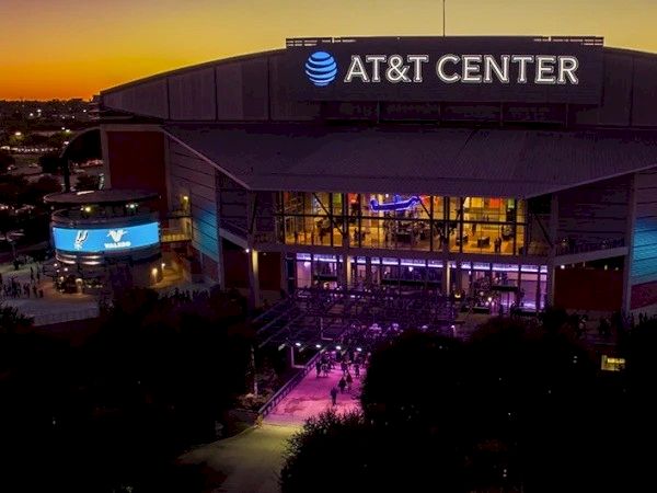 AT&T Center arena at dusk, illuminated entrance, sign lighting, football/hockey venue with surrounding trees and walkway.