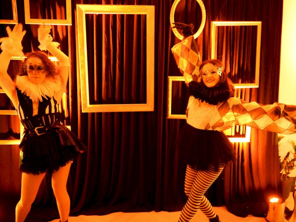 Two performers in vintage cabaret outfits pose playfully among geometric frames and warm fairy lights, in a retro stage set.