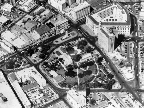 Aerial view of a city block with roads, mid-rise buildings, and a central park-like area featuring winding paths and greenery.