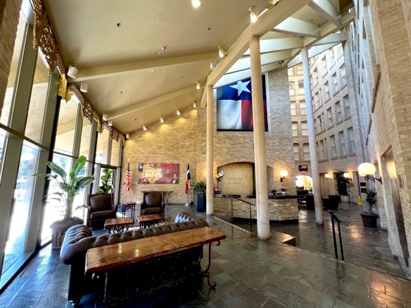 A stylish hotel lobby with high ceilings, stone walls, a Texas flag hanging above, modern seating, plants, and warm lighting.