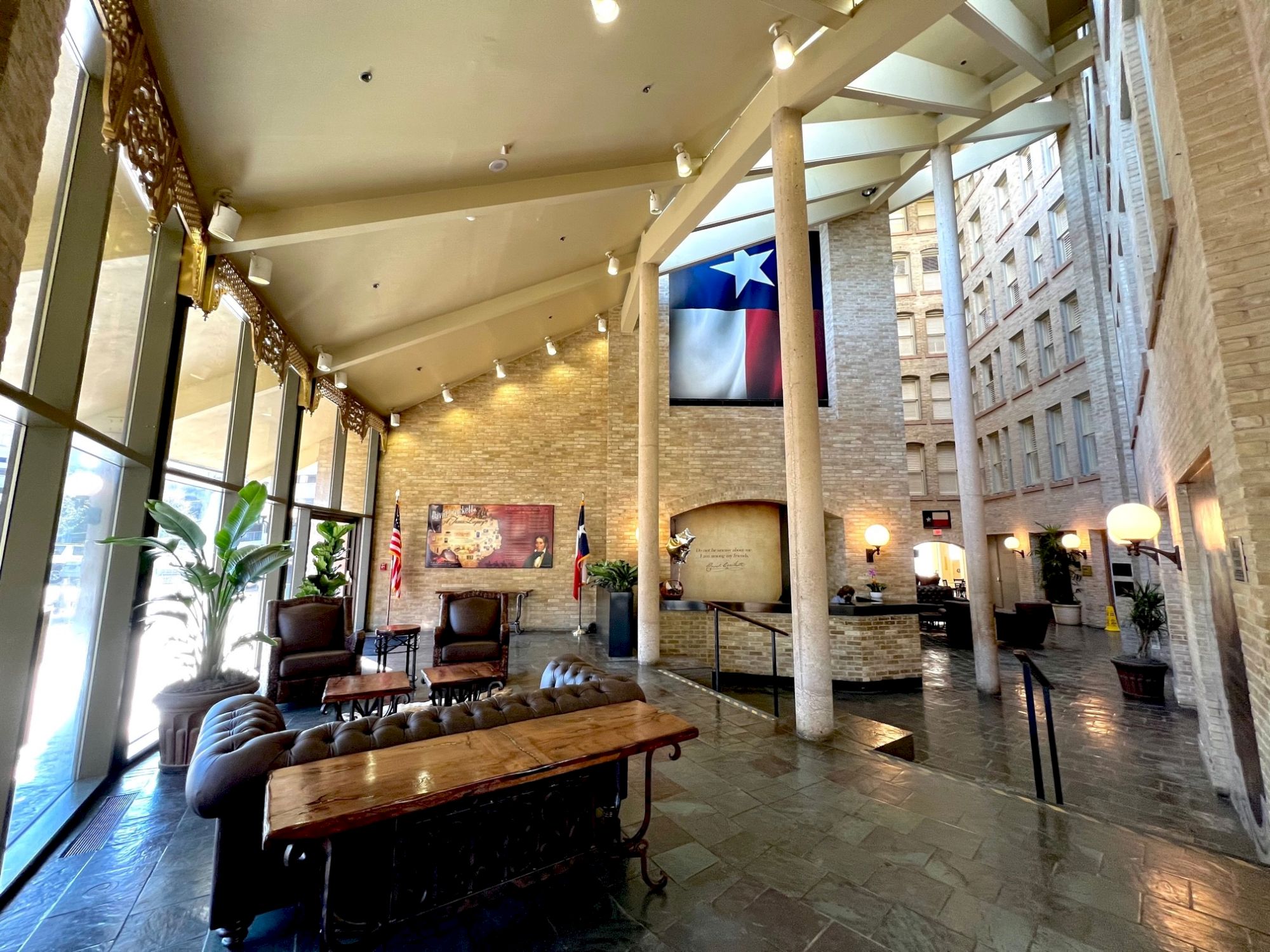 A stylish hotel lobby with stone walls, tall ceilings, string lights, and a Texas flag hanging; cozy seating, plants, and modern decor.
