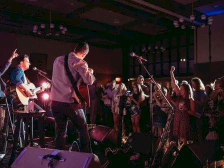 A live band performs on stage with singers and musicians holding guitars, brass, and microphones under colorful lights.