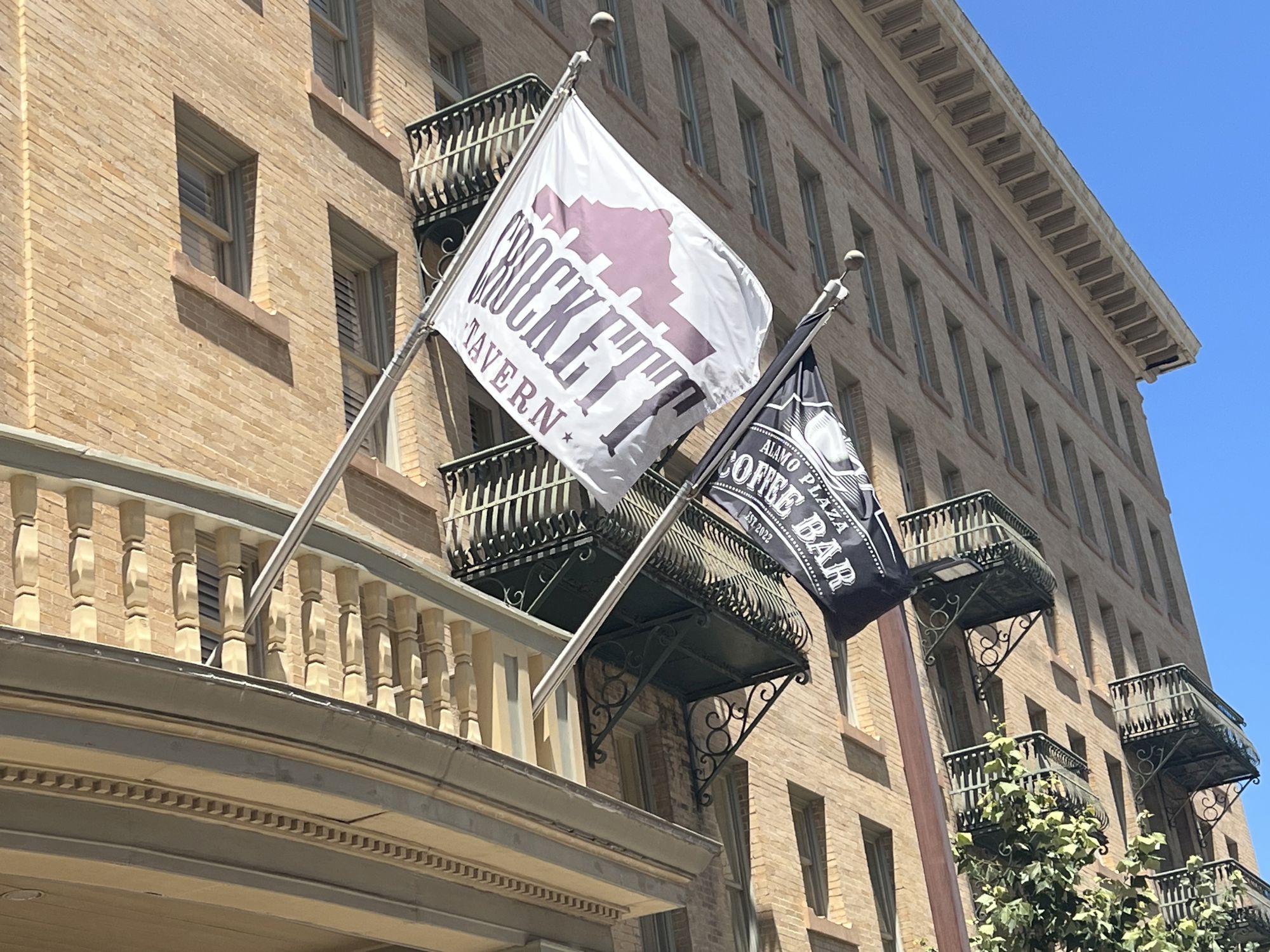 A historic brick building with two large banners, including an &ldquo;Alamo&rdquo; cafe flag, hanging between balconies in the sunlight.