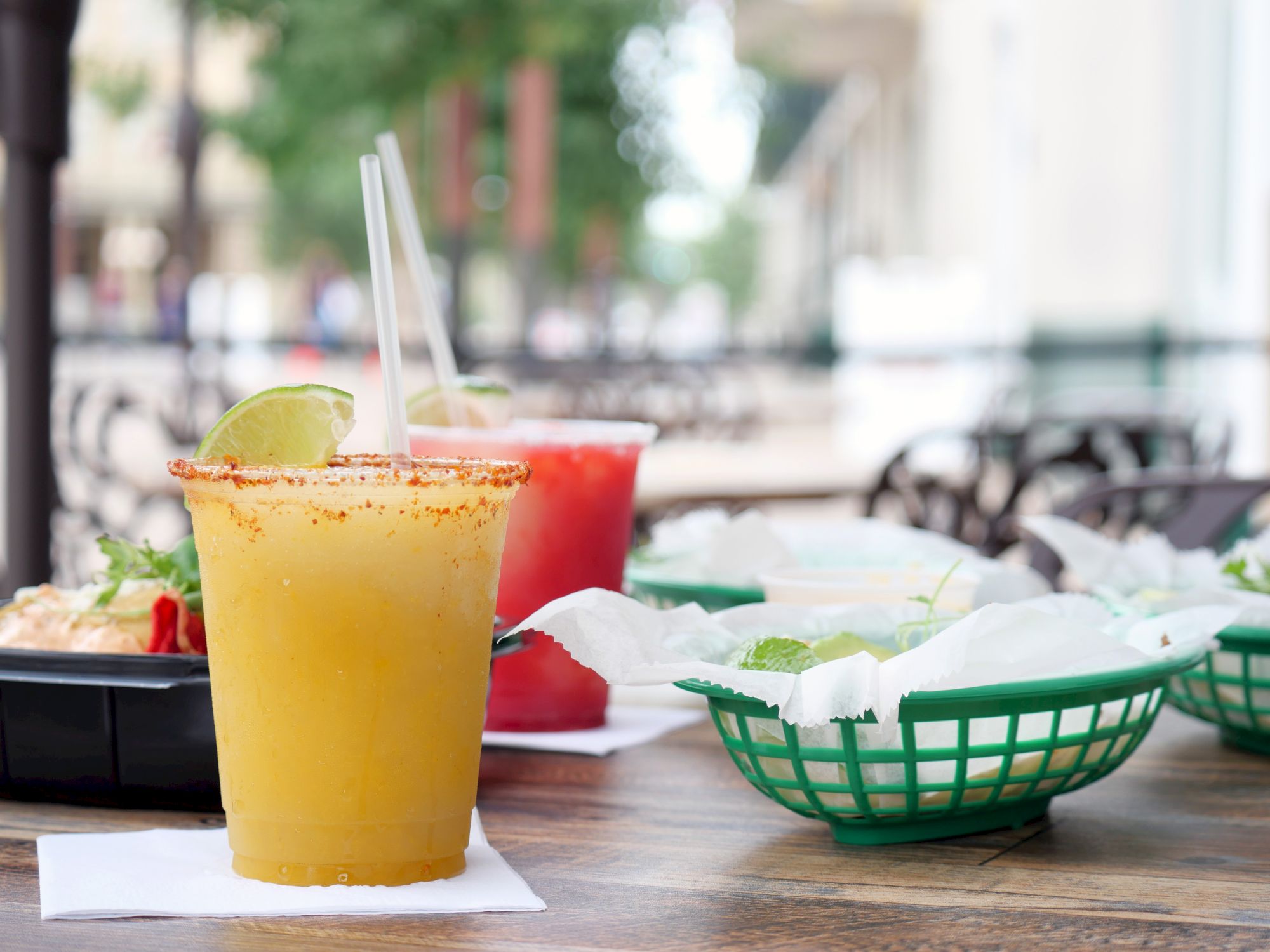A lime margarita-style drink with two straws sits on a table at an outdoor caf&eacute;, with another red drink blurred behind.