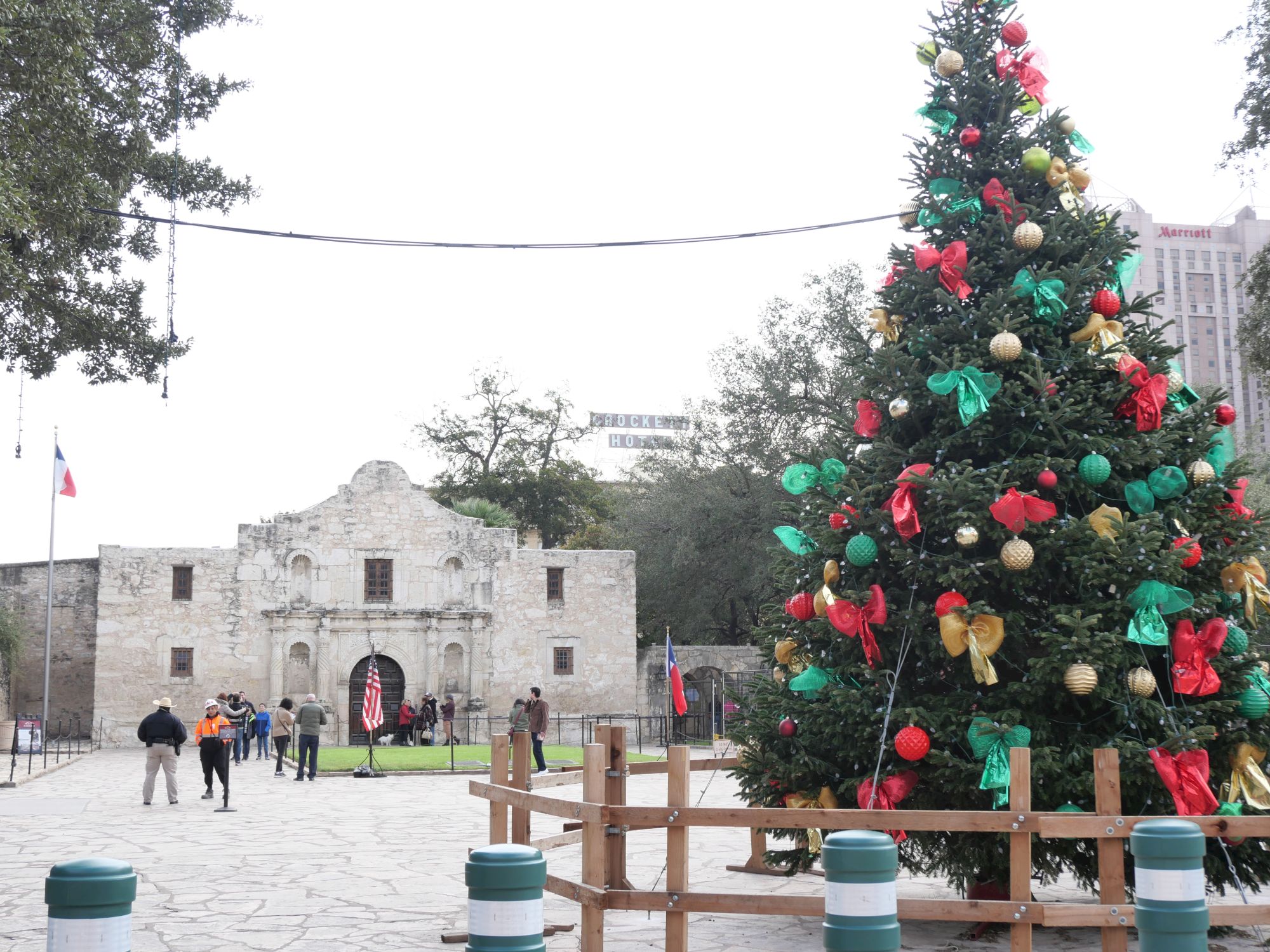 A huge decorated Christmas tree with red and green ornaments and bows stands outside a historic stone building as people walk by.