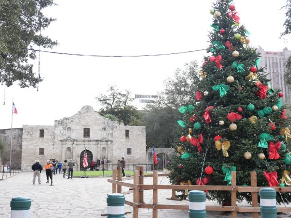 A huge Christmas tree covered in colorful ornaments and ribbons stands outside a stone building in a city square.